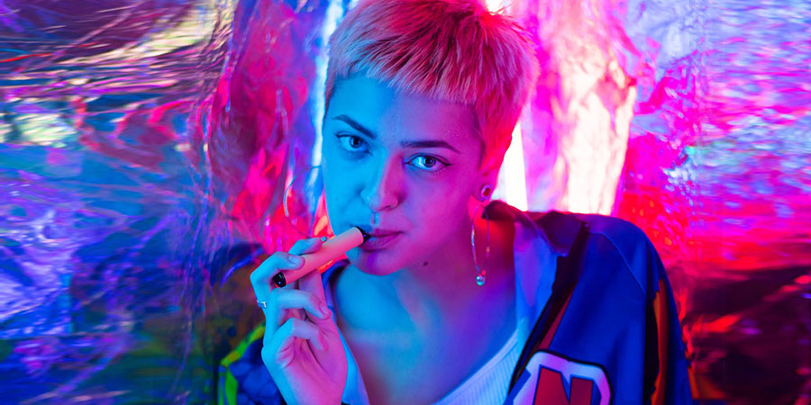 woman with short blonde hair wearing blue jacket holding a vape device with her hand and between her lips